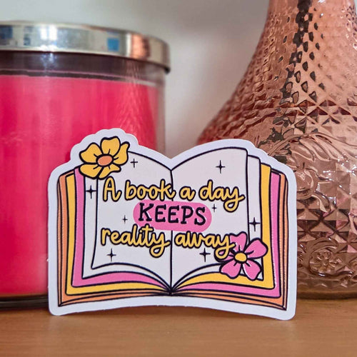 A die-cut sticker shaped like an open book with the phrase 'A book a day KEEPS reality away' in yellow script, placed on a wooden surface in front of a pink candle and a copper-toned textured vase.