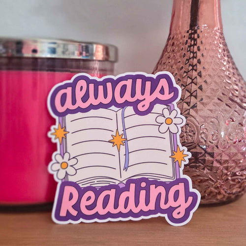 A die-cut sticker with the words 'always Reading' in pink and purple lettering, surrounding an open book illustration with flowers and stars, displayed on a wooden surface between decorative pink items.
