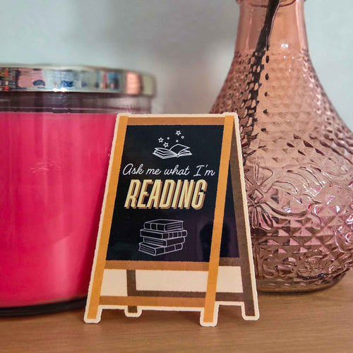 sticker of a small wooden sign with 'Ask me what I'm reading' text next to a pink candle and decorative vase.