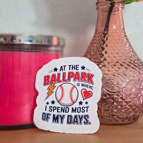Sticker with a baseball design and text on a surface with a pink container and vase in the background.