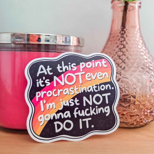 A die-cut sticker with a humorous procrastination quote, displayed on a wooden surface next to a pink candle and a textured rose gold vase.
