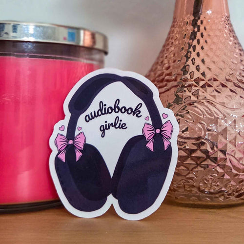 A clear die-cut sticker shaped like over-ear headphones with pink bows and the words 'audiobook girlie' in black script, displayed on a wooden surface between a pink candle and a textured rose-gold glass bottle.