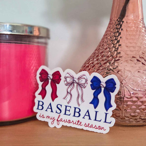 Sticker with decorative bows and text 'BASEBALL is my favorite season' on a surface with a pink candle and decorative vase.