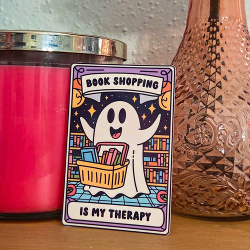 A rectangular sticker featuring a cartoon ghost holding a book basket with the text 'BOOK SHOPPING IS MY THERAPY', displayed on a wooden surface next to a pink candle and a patterned bottle.