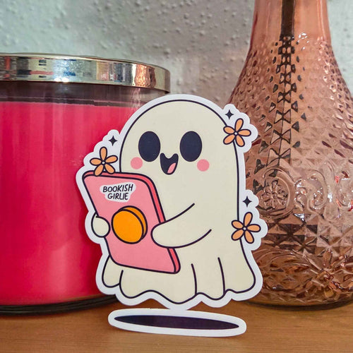 A die-cut sticker featuring a cute cartoon ghost holding a pink device that reads 'BOOKISH GIRLIE', displayed on a wooden surface between a pink candle and a textured copper-toned bottle.