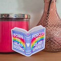 Sticker with rainbow design and text 'Born to read books, Forced to read emails' on a surface with a candle and vase.