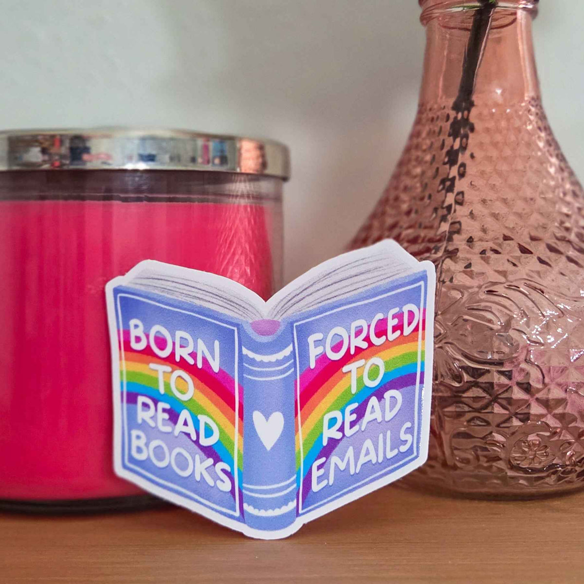 Sticker with rainbow design and text 'Born to read books, Forced to read emails' on a surface with a candle and vase.