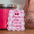A die-cut sticker with a pink and white gingham pattern, red cherries, and the text 'Buy me books & tell me I'm pretty' in red script, displayed on a wooden surface next to a pink candle and a copper-colored textured bottle.