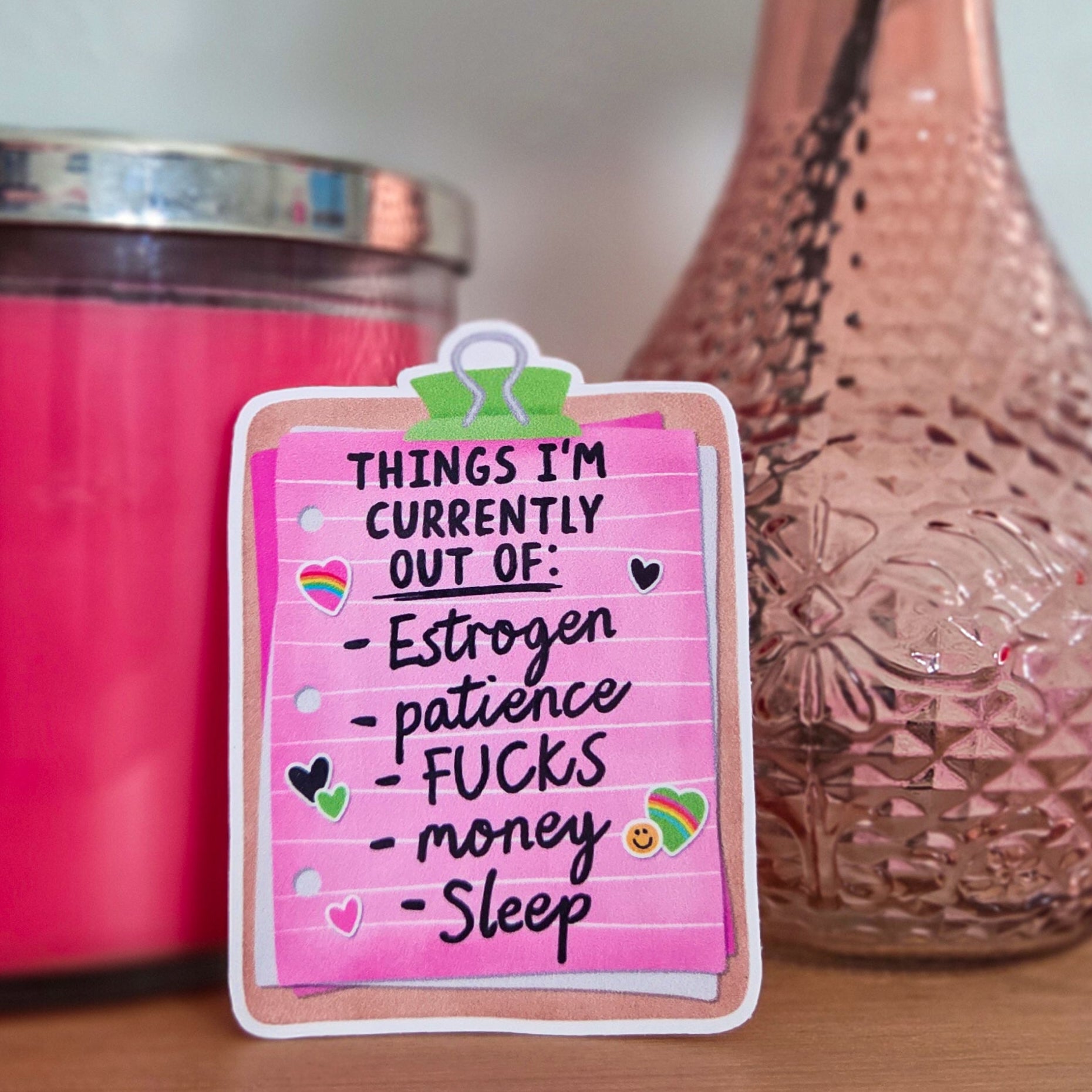 Pink card with text on a wooden surface next to a pink candle and vase with flowers.