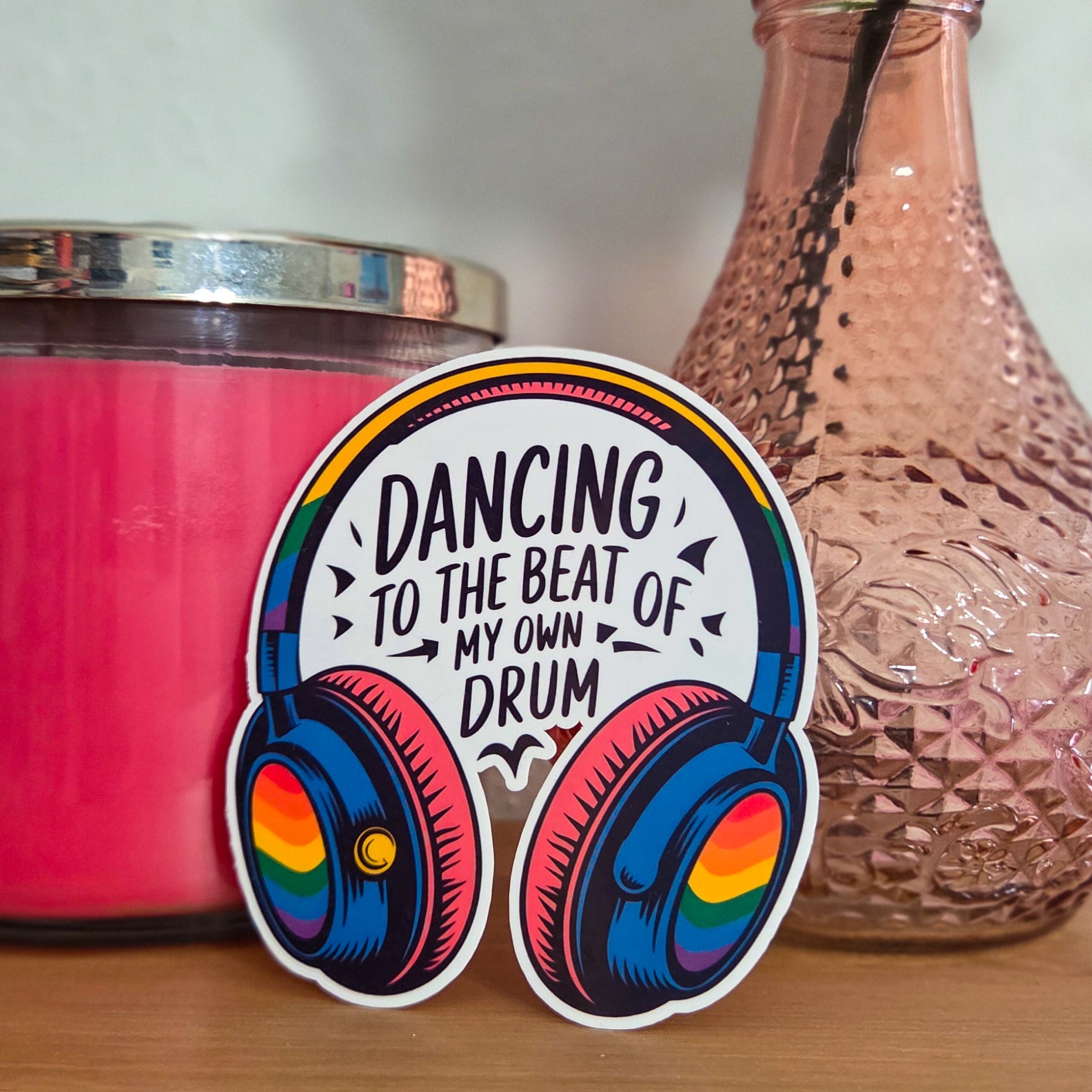 Sticker with headphones design and text 'Dancing to the beat of my own drum' on a wooden surface with a pink candle and vase in the background.