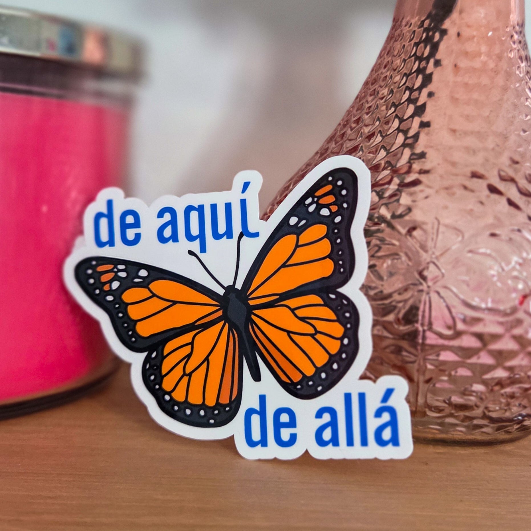 Butterfly sticker with text 'de aqui de allá' on a wooden surface with a pink vase and candle.