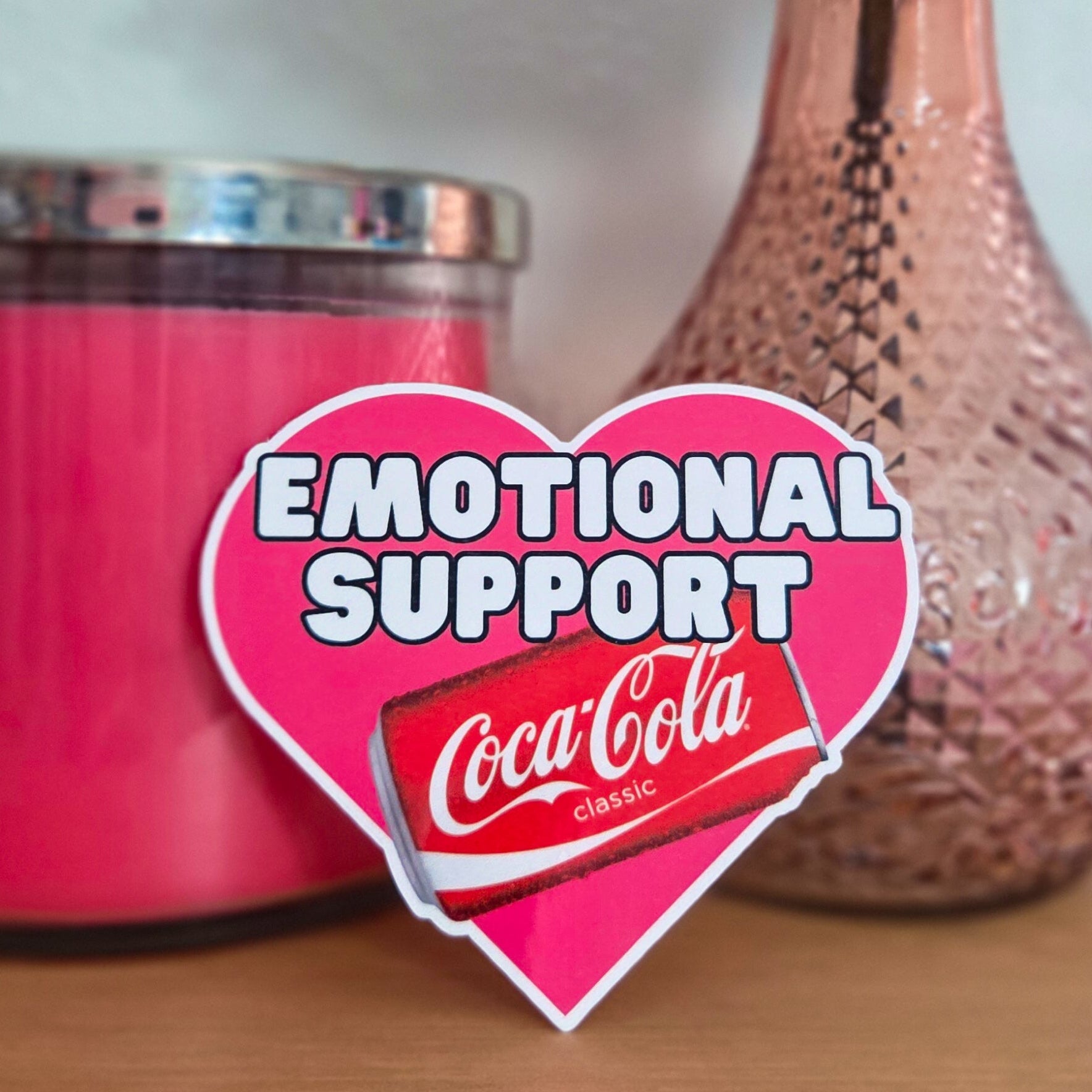 Pink candle, vase with peach rose, and Emotional Support Coke heart-shaped sticker on a wooden surface.