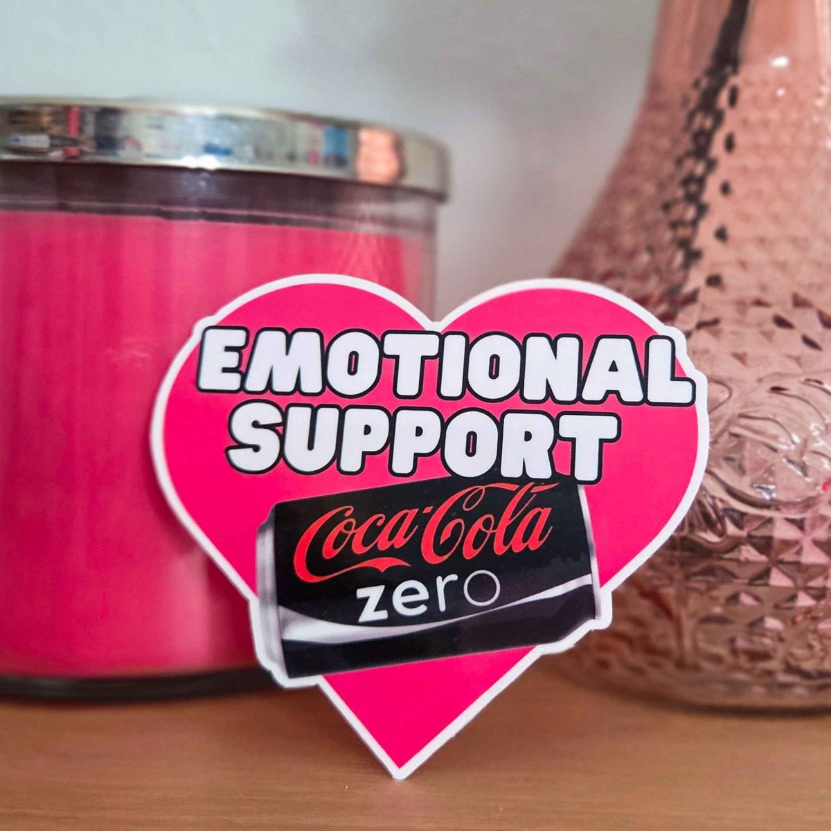Heart-shaped 'Emotional Support' sticker with Coke Zero can in front of pink candle and decorative vase.