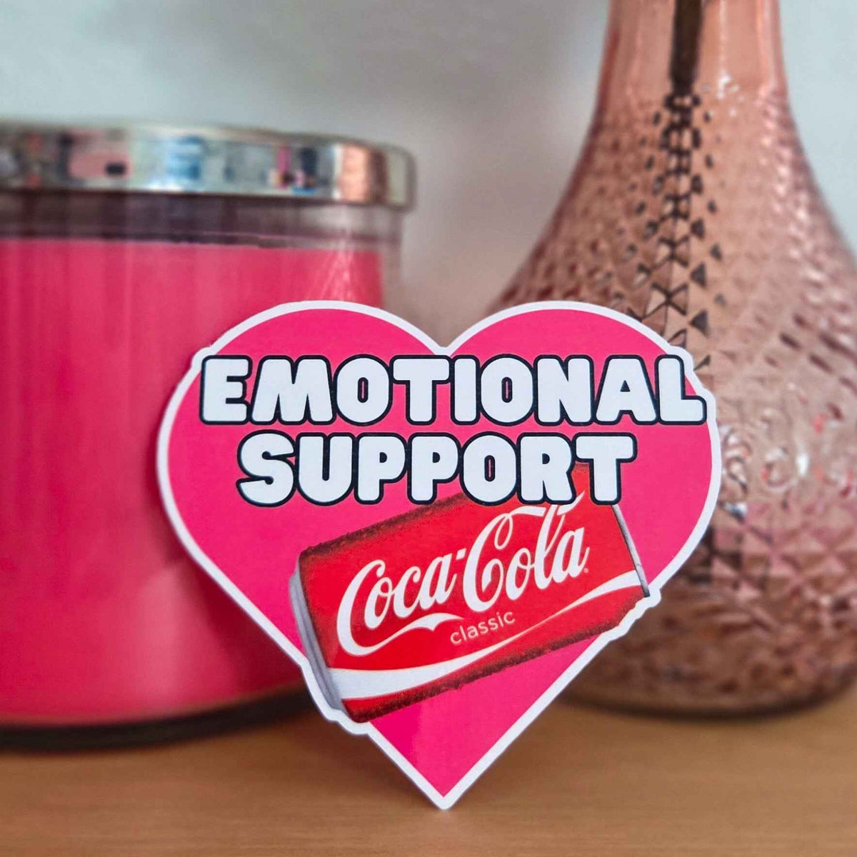 Heart-shaped 'Emotional Support' sticker with Coke can in front of pink candle and decorative vase.