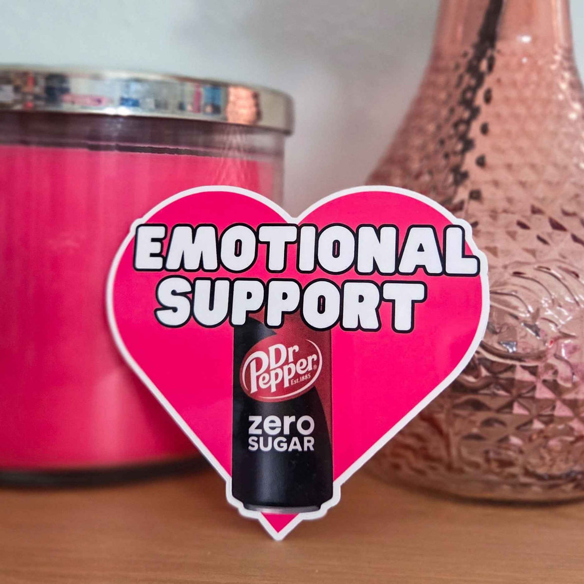 Heart-shaped 'Emotional Support' sticker with Dr Pepper Zero can in front of pink candle and decorative vase.