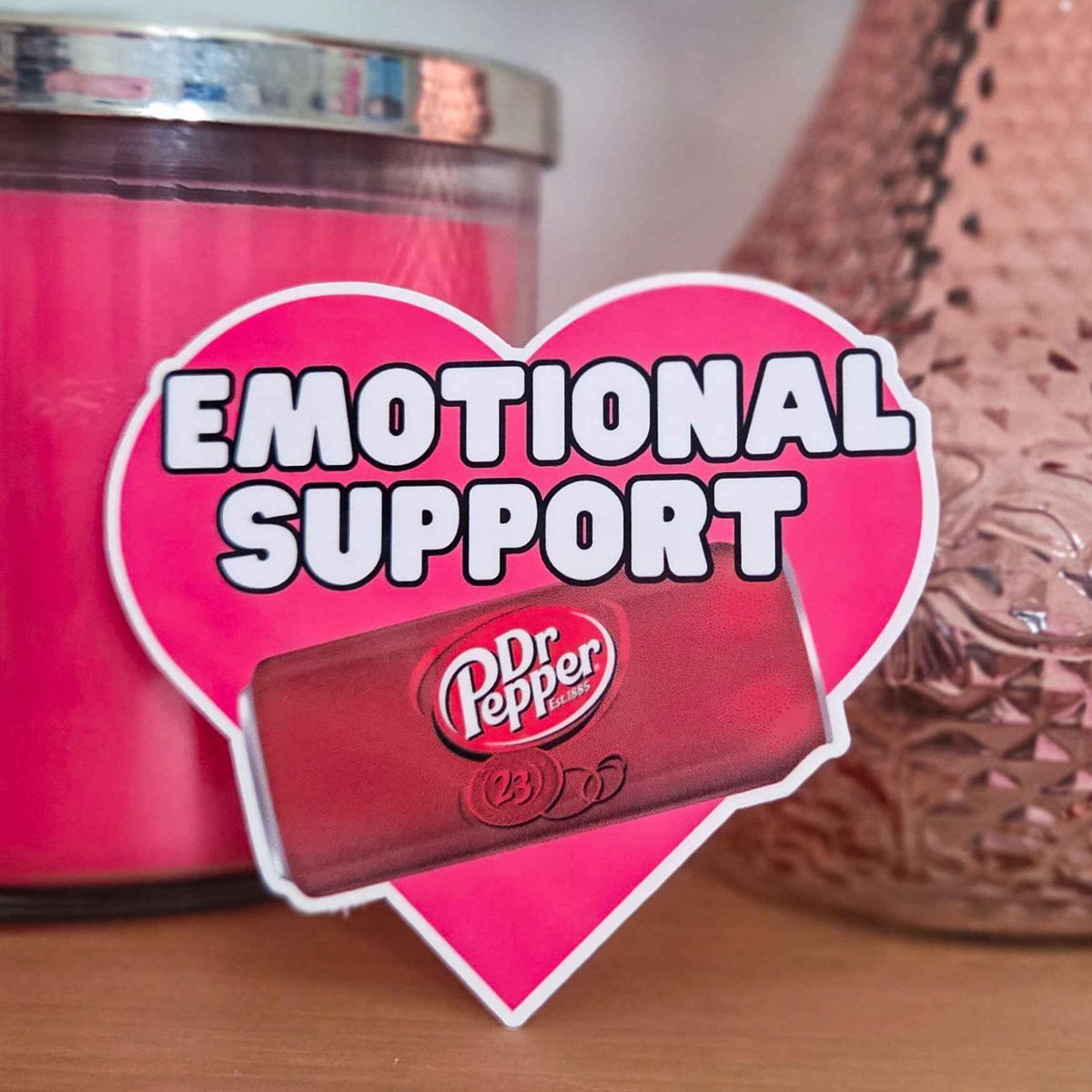 Heart-shaped 'Emotional Support' sticker with Dr Pepper can in front of pink candle and decorative vase.