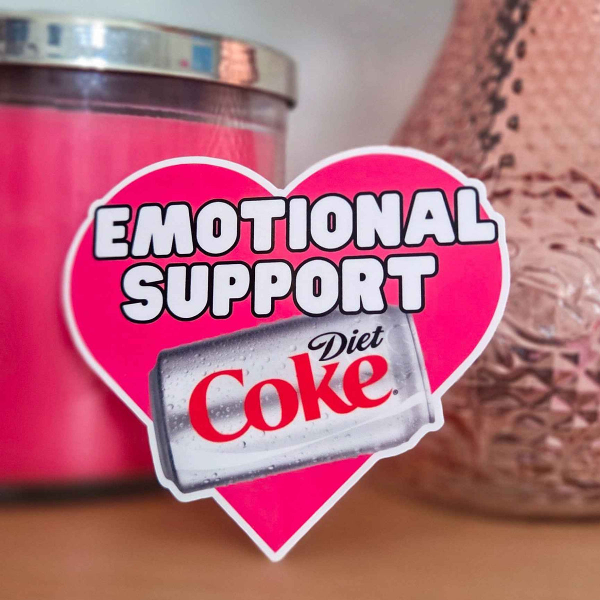 Heart-shaped 'Emotional Support' sticker with Diet Coke can in front of pink candle and decorative vase.