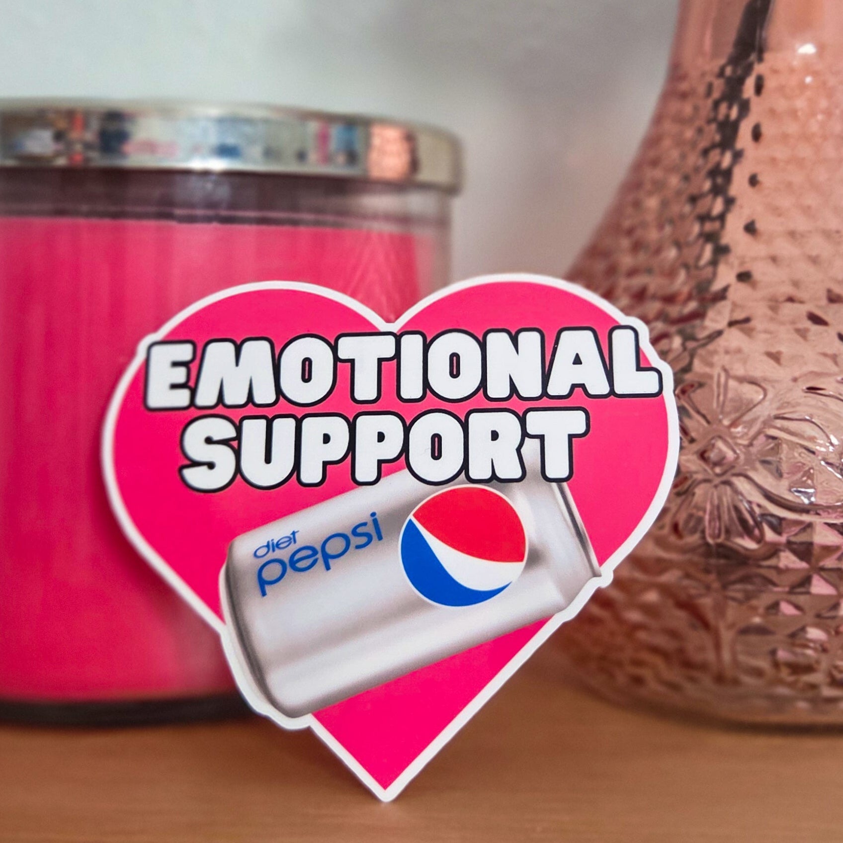 Pink candle and a vase with a heart-shaped 'Emotional Support' sticker featuring a Diet Pepsi logo on a wooden surface.