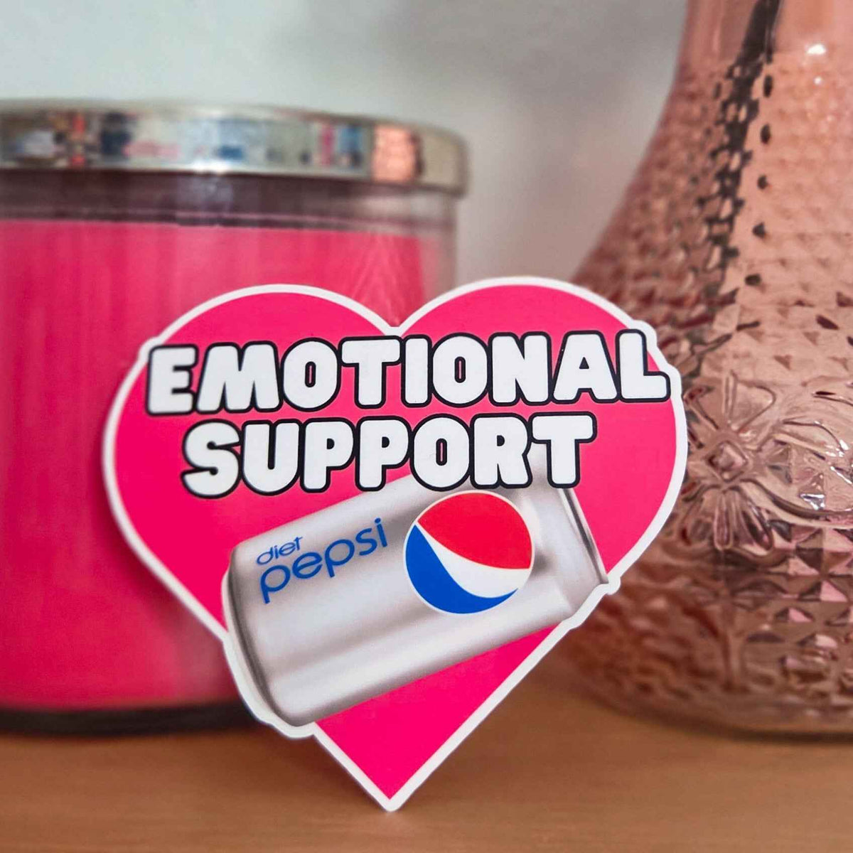 Heart-shaped 'Emotional Support' sticker with Diet Pepsi can in front of pink candle and decorative vase.