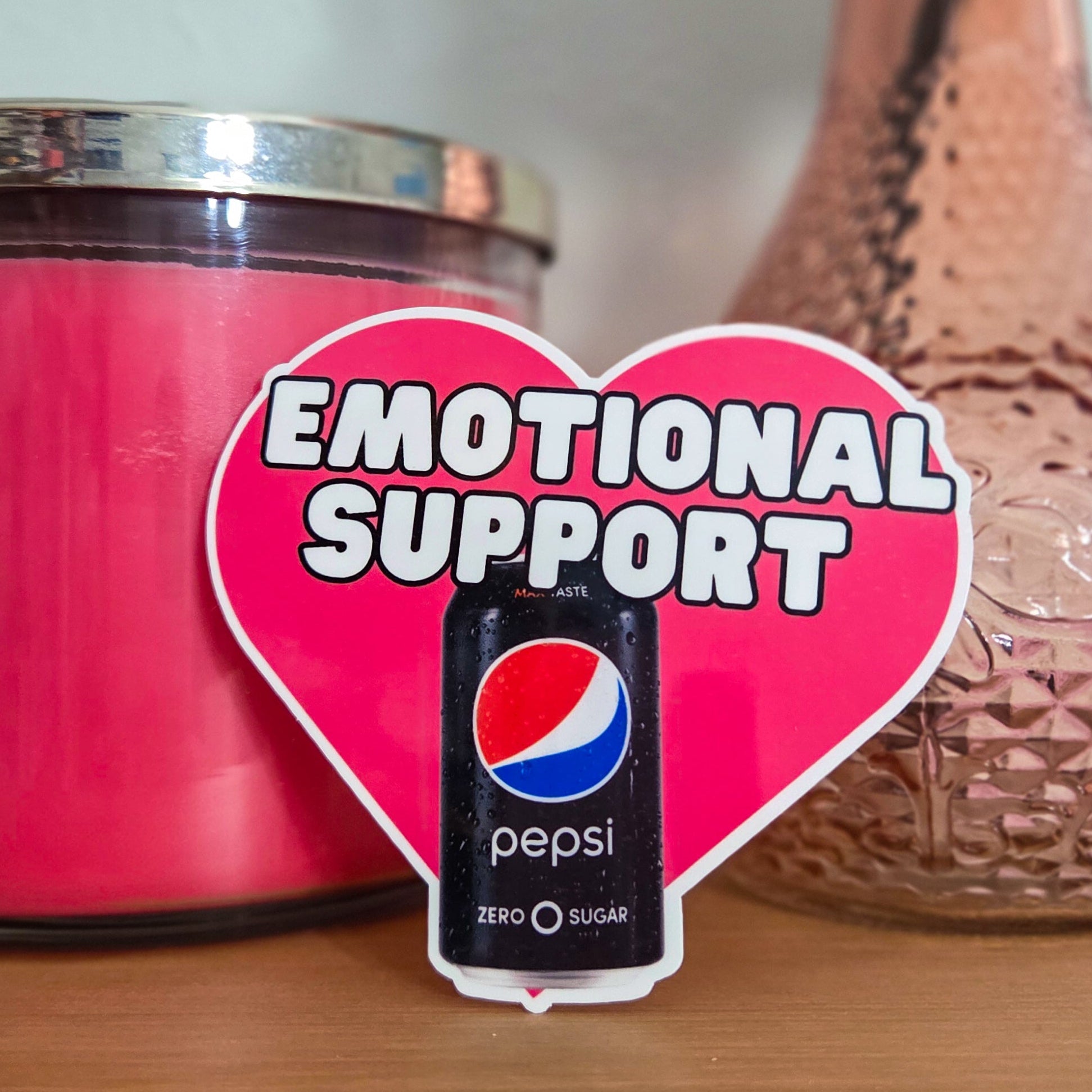 Heart-shaped 'Emotional Support' sticker with Pepsi logo next to a pink candle and vase on a wooden surface.