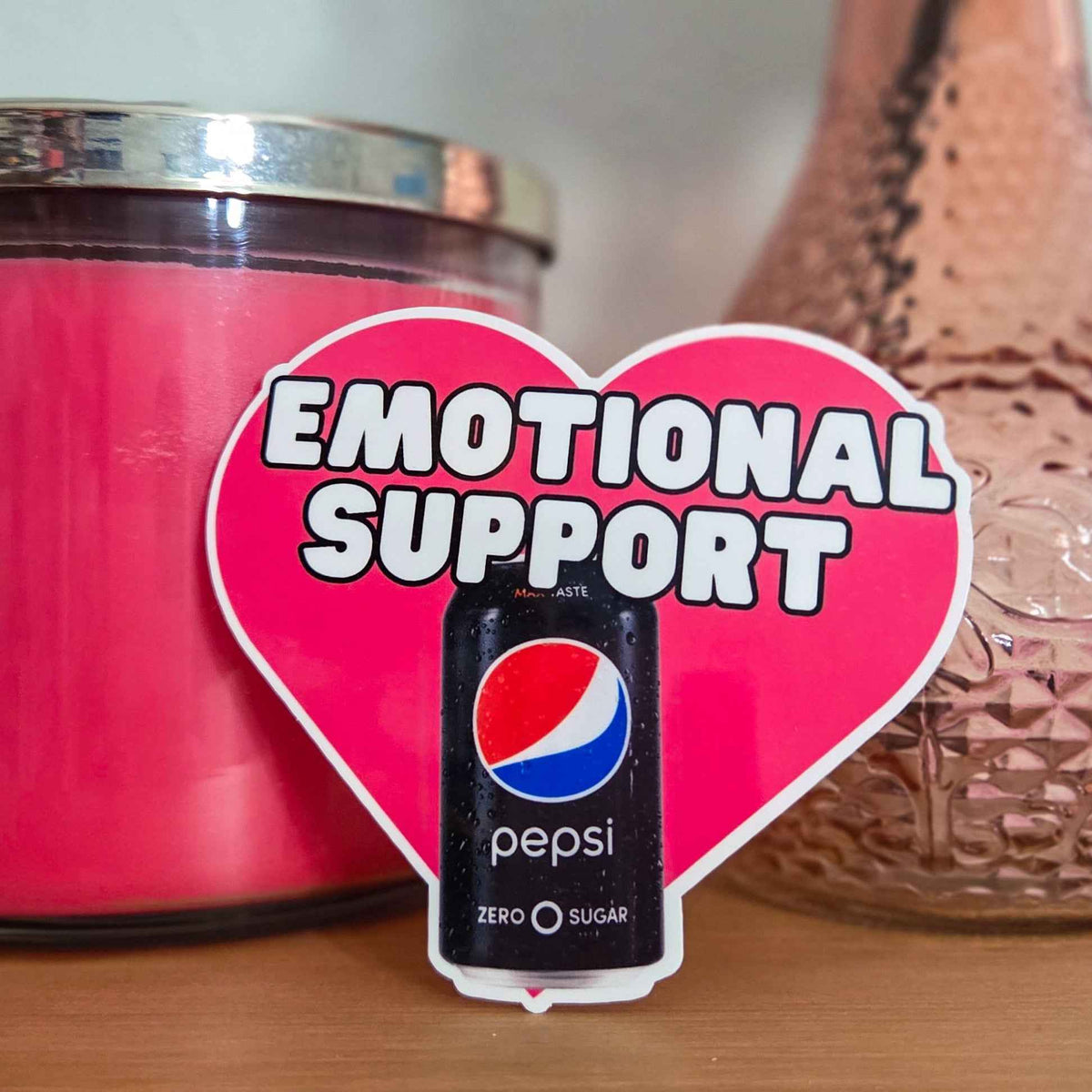 Heart-shaped 'Emotional Support' sticker with Pepsi Zero can in front of pink candle and decorative vase.