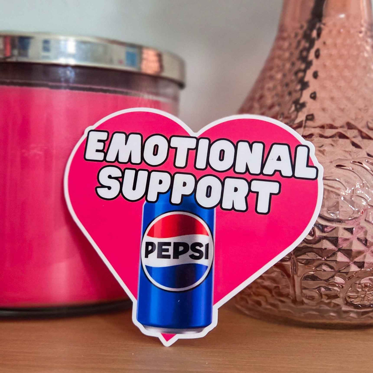 Heart-shaped 'Emotional Support' sticker with Pepsi can in front of pink candle and decorative vase.
