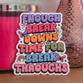 A die-cut sticker with a colorful design that reads ‘Enough Breakdowns Time for Breakthroughs ' resting on a wooden surface.