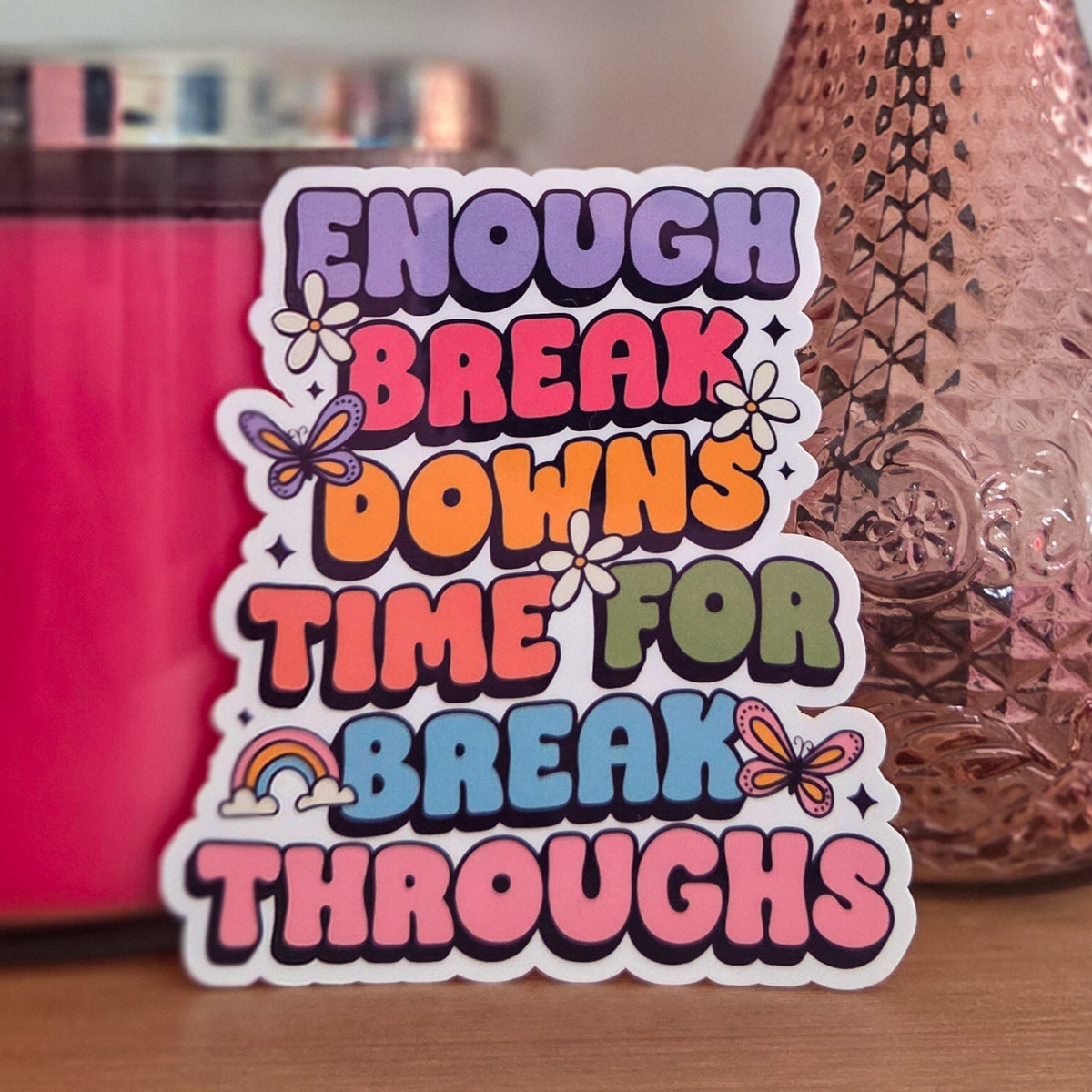 A die-cut sticker with a colorful design that reads ‘Enough Breakdowns Time for Breakthroughs ' resting on a wooden surface.