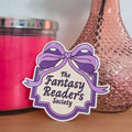 Pink candle, pink vase with a rose, and 'The Fantasy Reader's Society' sticker on a wooden surface.