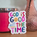 Colorful 'God is Good all the Time' sticker on a wooden surface with a pink candle and vase in the background.