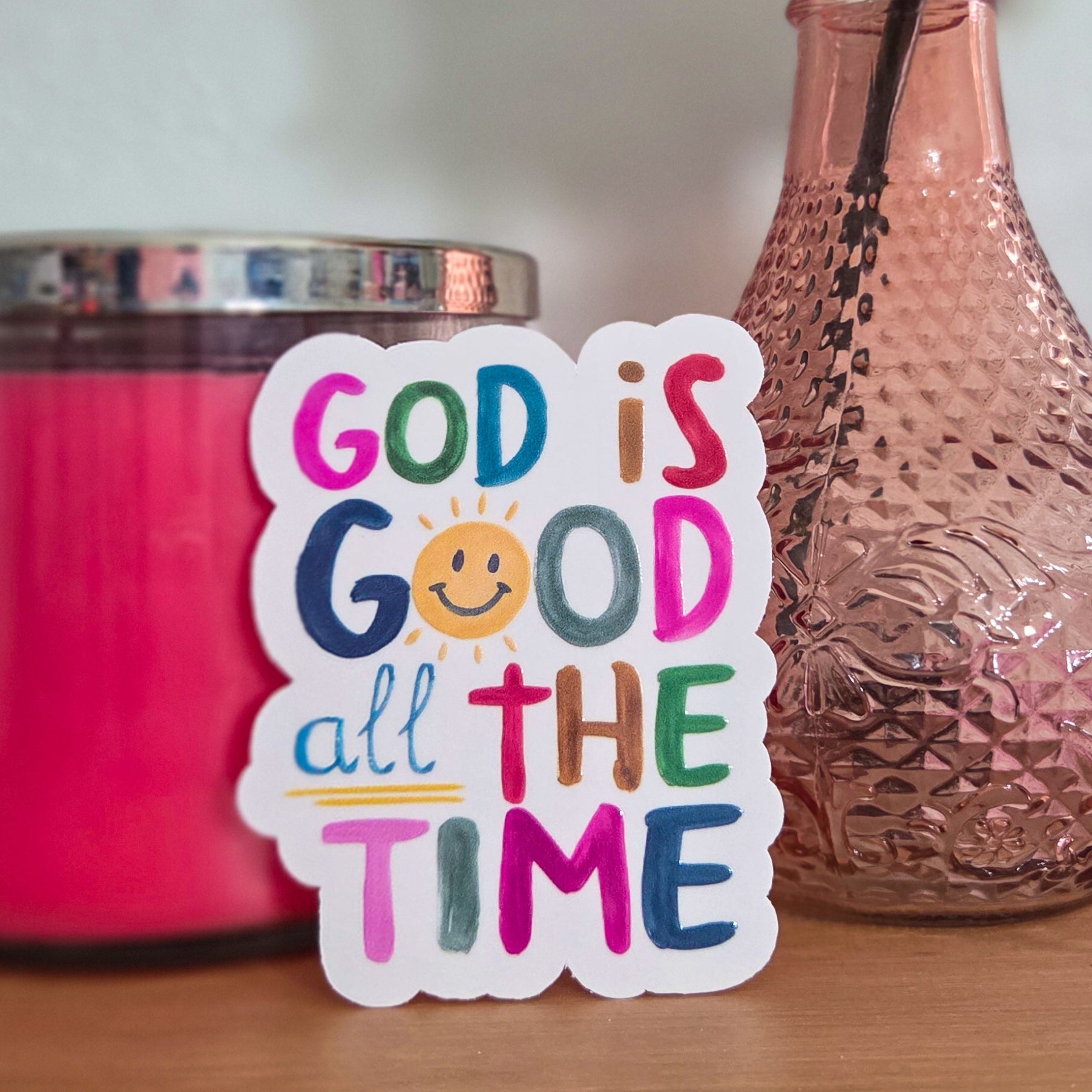 Colorful 'God is Good all the Time' sticker on a wooden surface with a pink candle and vase in the background.