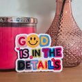 Colorful sticker with 'God is in the details' text next to a pink candle and vase with flowers on a wooden surface.