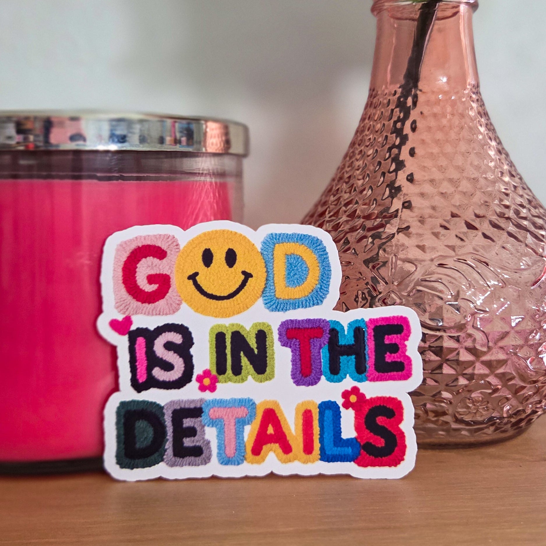 Colorful sticker with 'God is in the details' text next to a pink candle and vase with flowers on a wooden surface.