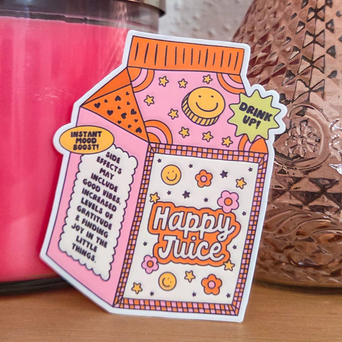 A die-cut sticker with a colorful design that reads ‘Happy Juice' resting on a wooden surface.