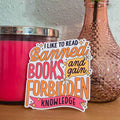 A die-cut sticker with the quote 'I LIKE TO READ Banned BOOKS and gain FORBIDDEN KNOWLEDGE' in colorful lettering, styled on a wooden surface next to a pink candle and a textured rose gold glass bottle.