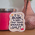 Sticker with humorous text on a surface with a pink candle and glass bottle in the background