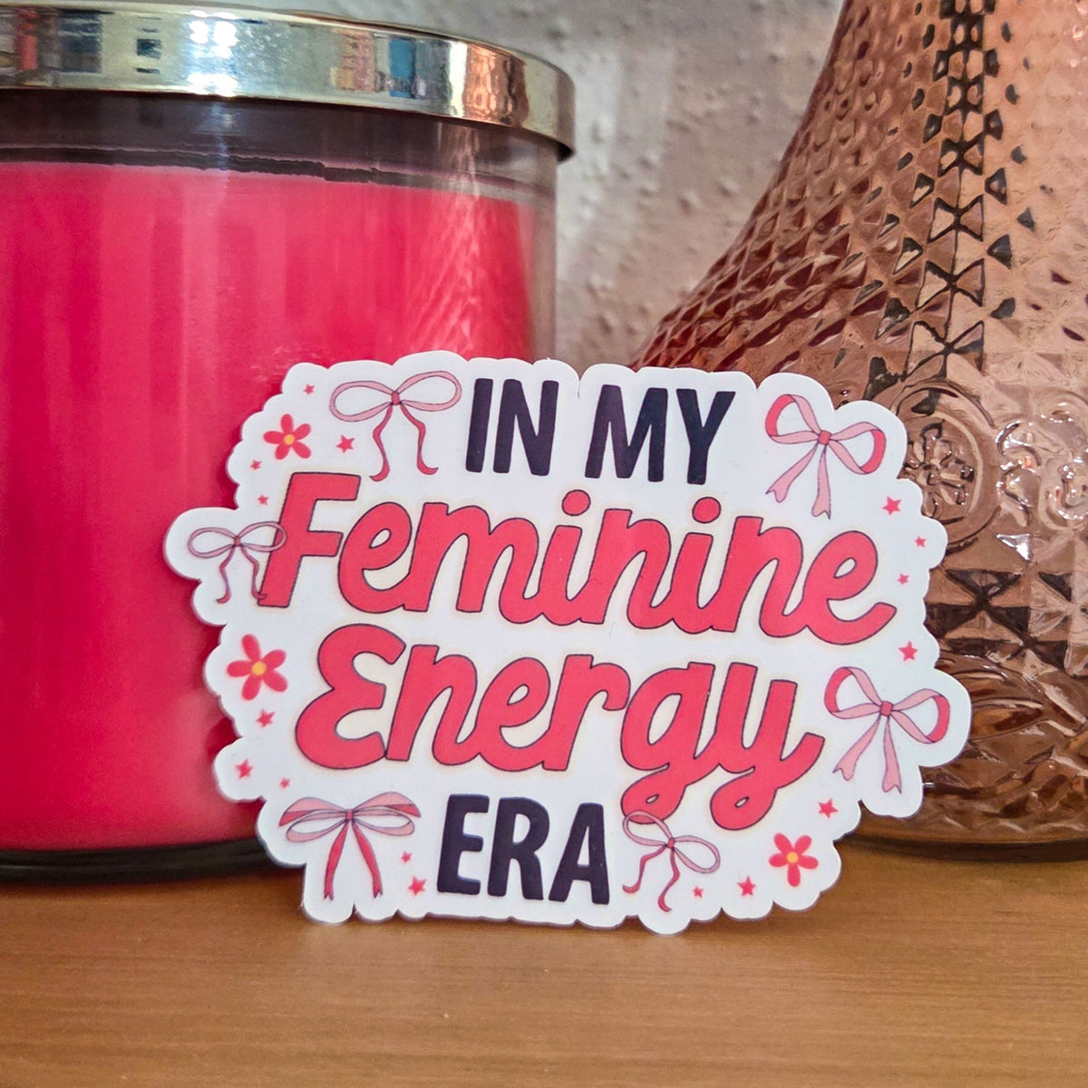 A die-cut sticker with a colorful design that reads ‘In My Feminine Energy Era' resting on a wooden surface.