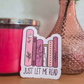 Sticker with book illustrations and 'Just Let Me Read' text on a wooden surface with a pink candle and decorative bottle in the background.
