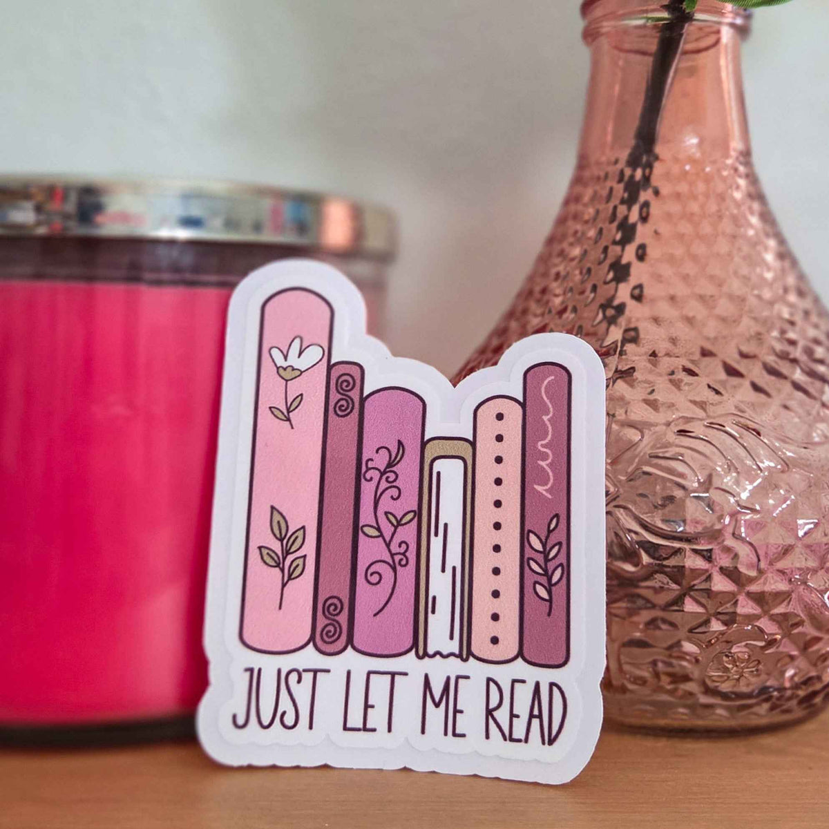 Sticker with book illustrations and 'Just Let Me Read' text on a wooden surface with a pink candle and decorative bottle in the background.