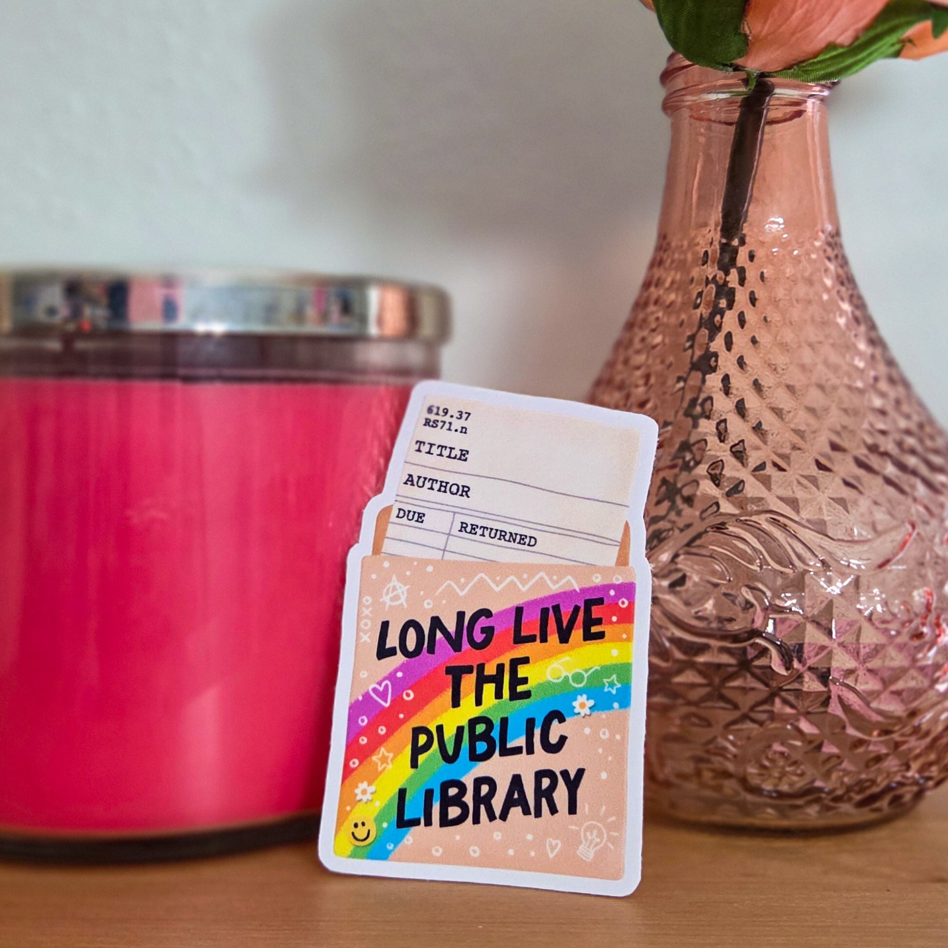 Card with 'Long Live the Public Library' text next to a pink candle and vase with a rose on a wooden surface.
