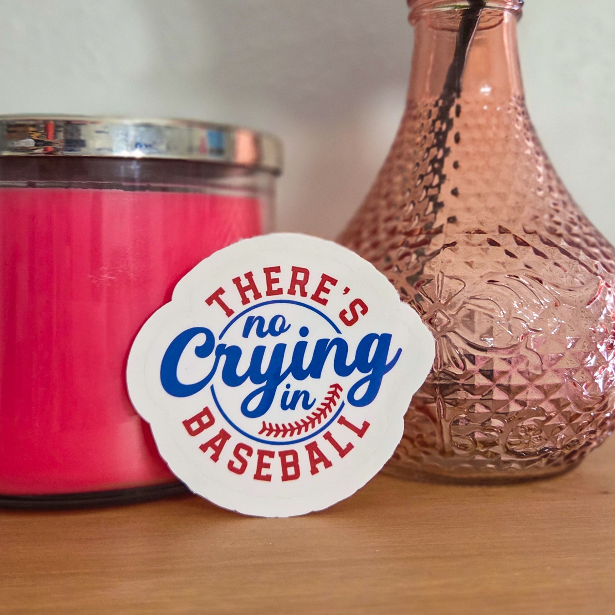 Pink candle and vase with a rose on a wooden surface, featuring a 'There's No Crying in Baseball' sticker.