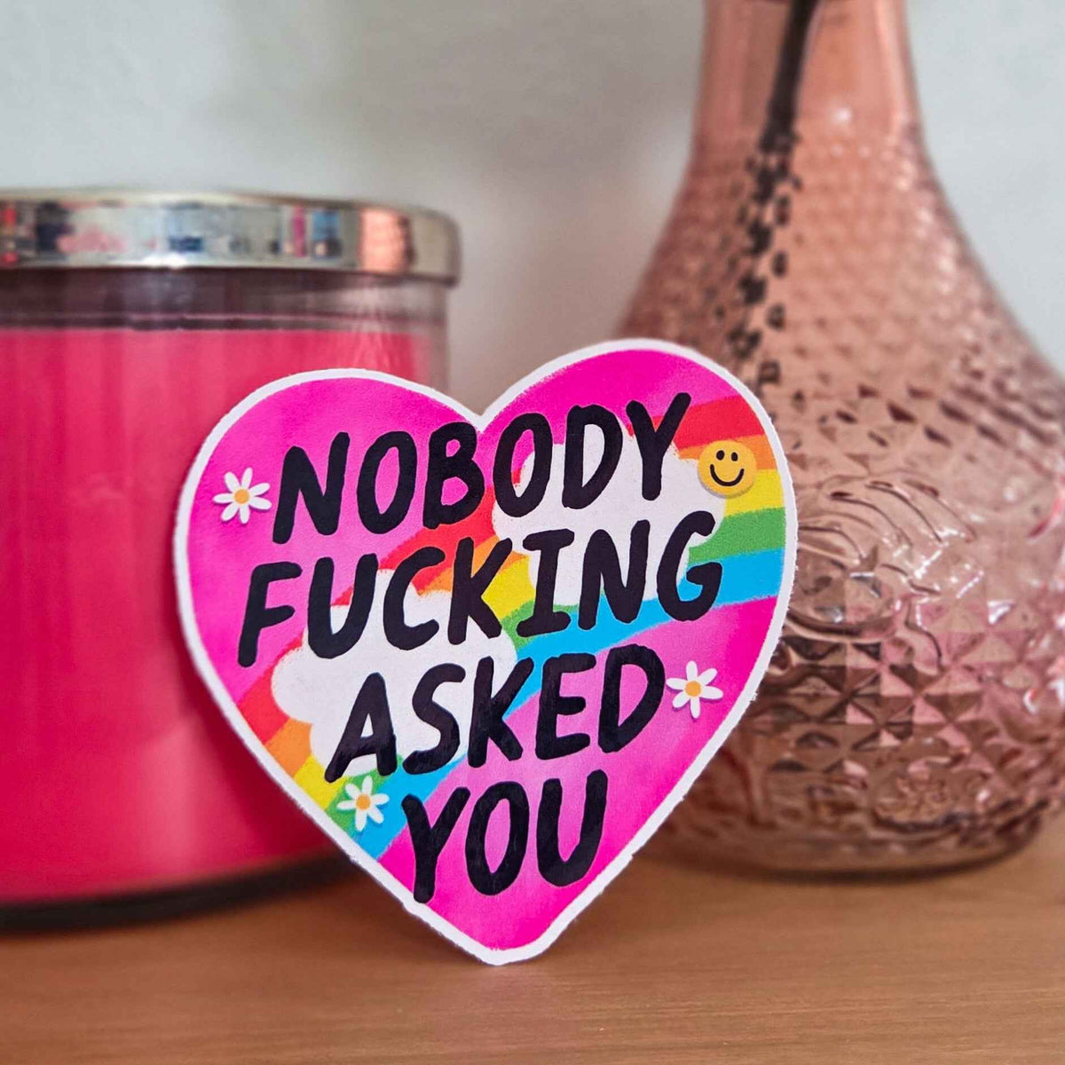 Heart-shaped sticker with colorful text on a surface with a pink candle and decorative vase.