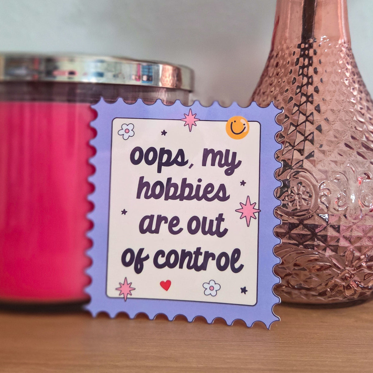 A die-cut sticker with a colorful design that reads ‘Oops, My Hobbies Are Out of Control Sticker' resting on a wooden surface.