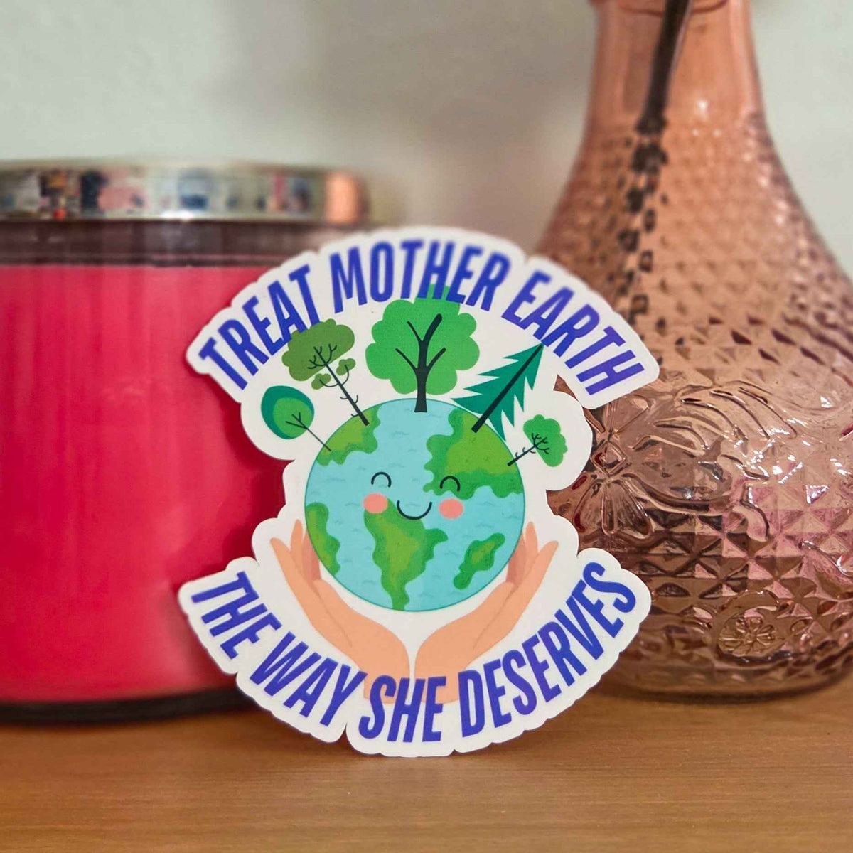 Sticker with 'Treat Mother Earth the way she deserves' text on a red candle and vase with flowers.