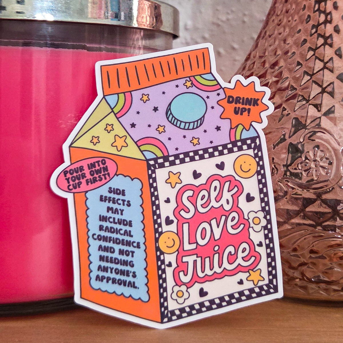 A die-cut sticker with a colorful design that reads ‘Self Love Juice' resting on a wooden surface.