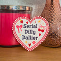A die-cut sticker with a colorful cute design that reads 'Serial Dilly Dallier’ resting on a wooden surface.