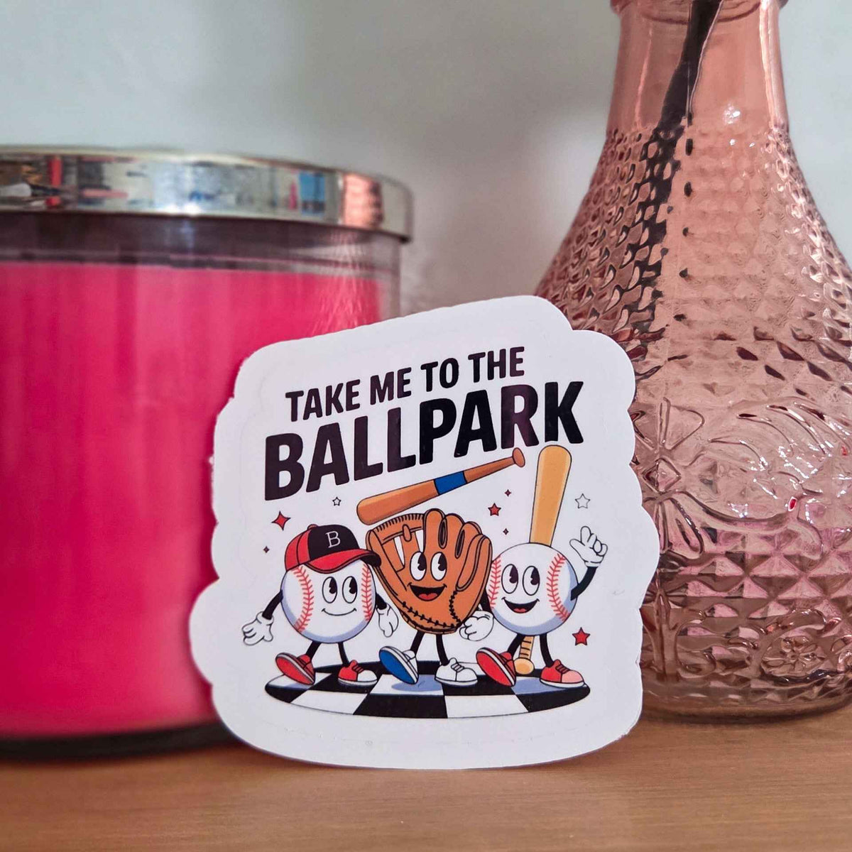 Sticker with cartoon characters and text 'Take me to the Ballpark' on a wooden surface with a pink candle and vase in the background.