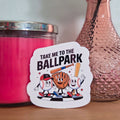 Sticker with cartoon characters and text 'Take me to the Ballpark' on a wooden surface with a pink candle and vase in the background.