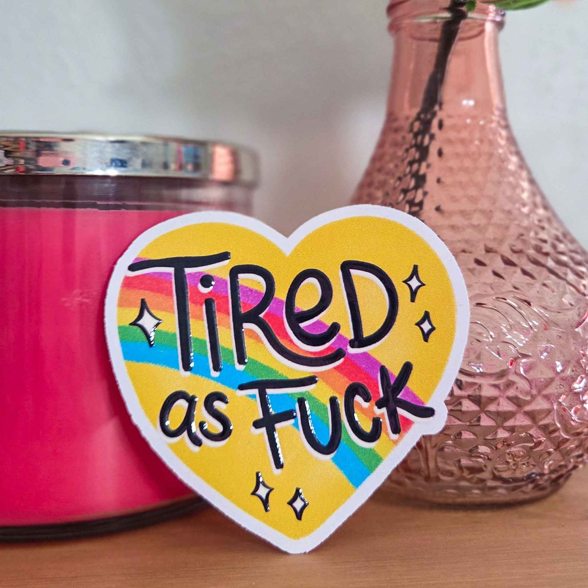 Heart-shaped sticker with 'Tired as Fuck' text next to a pink candle and decorative vase.