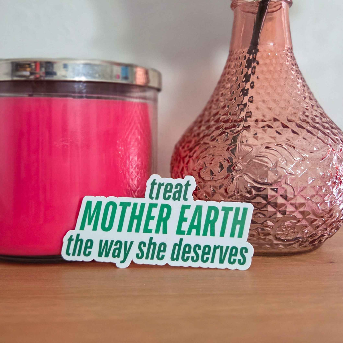 Pink candle and vase with a rose on a wooden surface, featuring a 'treat Mother Earth' sticker.
